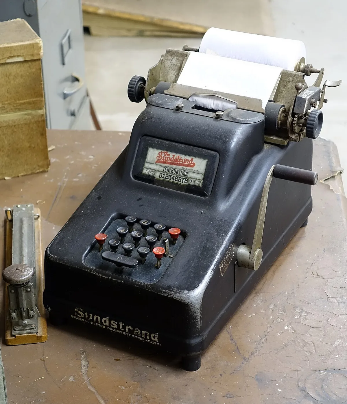 Zwarte mechanische telmachine met ronde toetsen en een hendel aan de zijkant, staand op een houten werkblad.