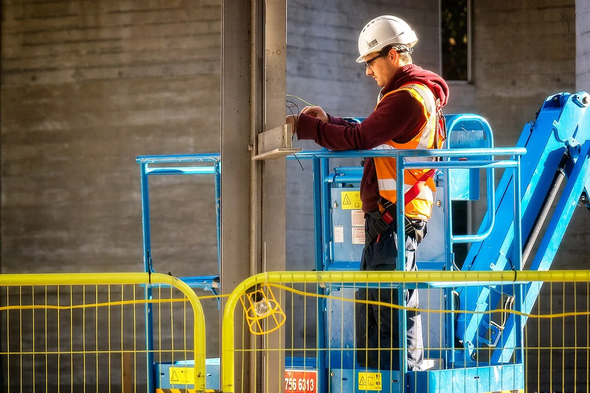 Elektricien met veiligheidshelm en oranje hesje op een blauw hoogwerkplatform, werkend aan bekabeling tegen het plafond van een lege bedrijfsruimte.