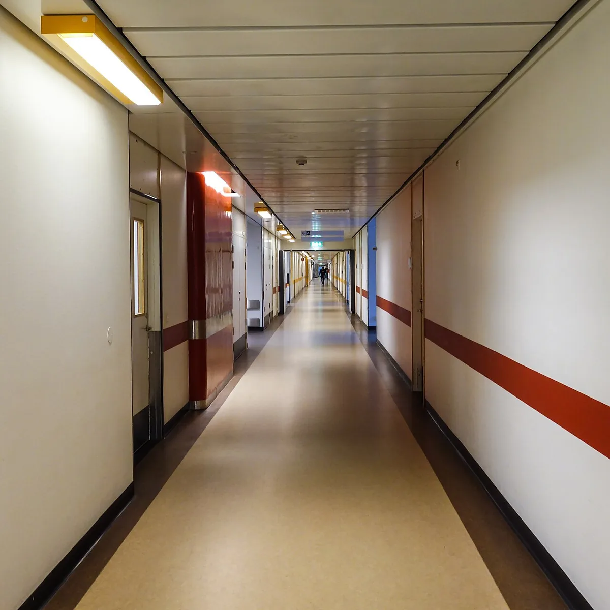 Lange ziekenhuisgang met lichte wanden, een rode accentstreep en een verlaagd plafond met tl-verlichting, in de verte loopt een persoon.