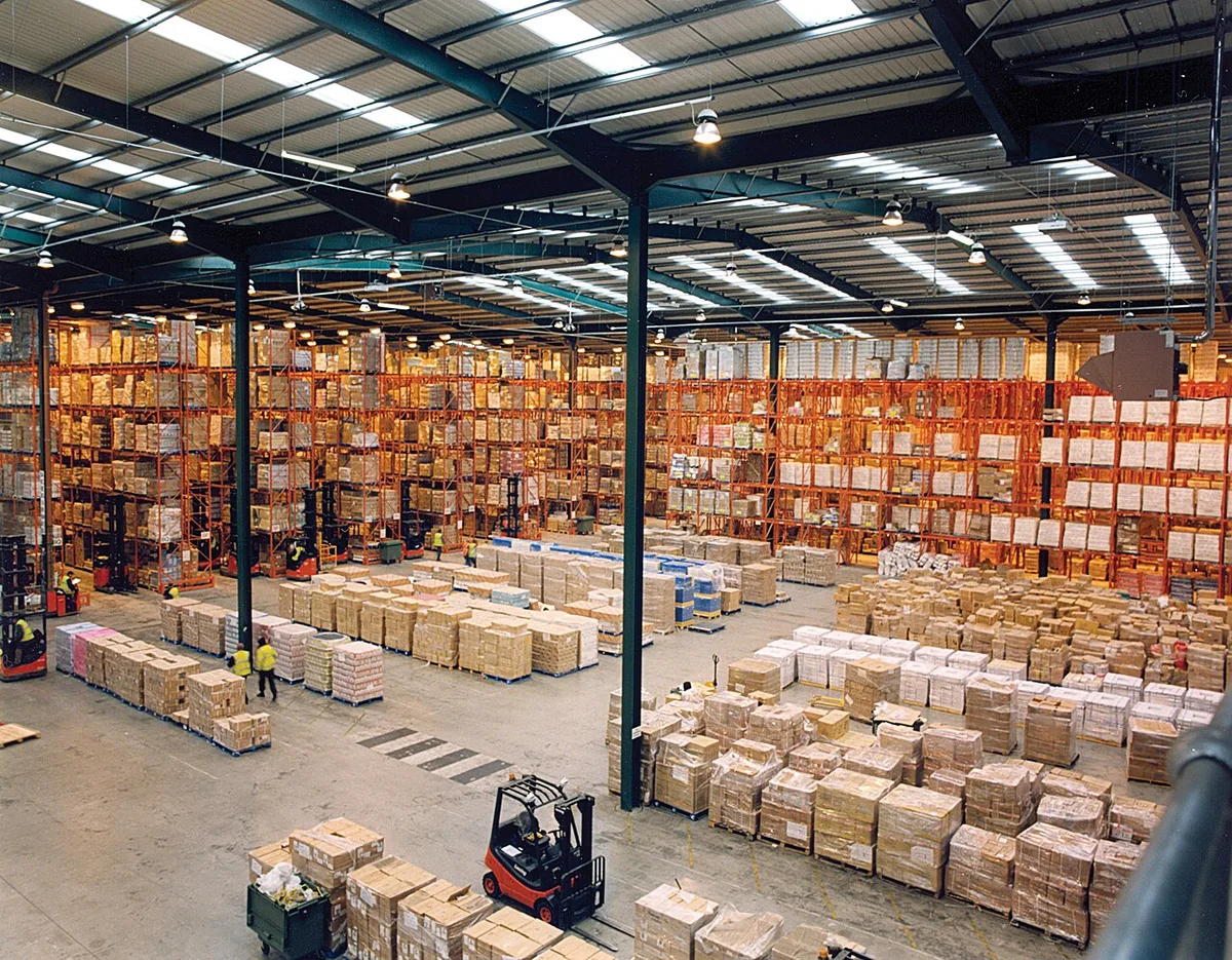 Overzicht van een groot magazijn met hoge stellingen vol dozen en pallets, een heftruck op de vloer en daglicht door de daklichten.