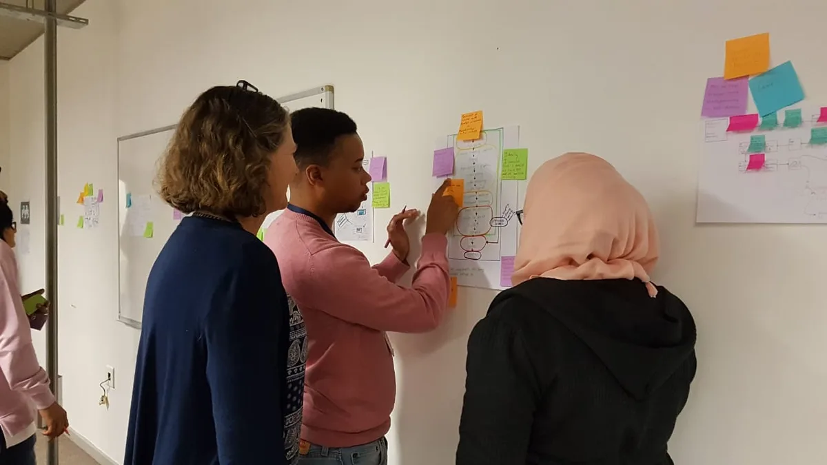Drie mensen staan voor een whiteboard met gekleurde plakbriefjes en schrijven met een stift een schema uit tijdens een werksessie.