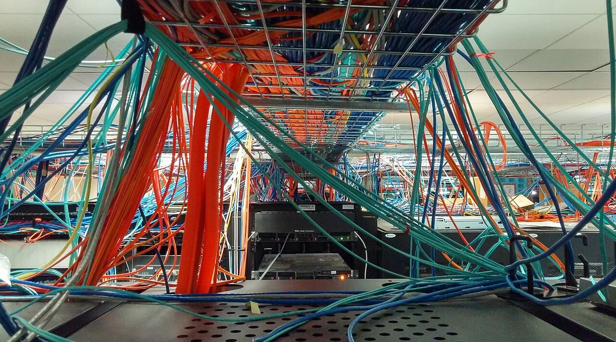 Dicht op elkaar liggende bundels oranje en turkooizen netwerkkabels onder een verhoogde computervloer, uitwaaierend naar meerdere kanten.